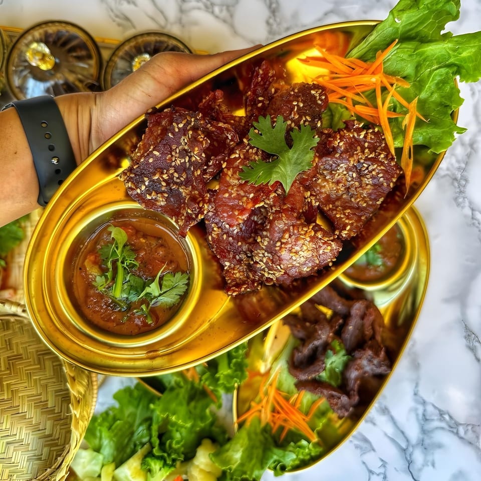 A hand holds a golden platter of So Saap's crispy Thai beef with sesame seeds, sauce, and greens.